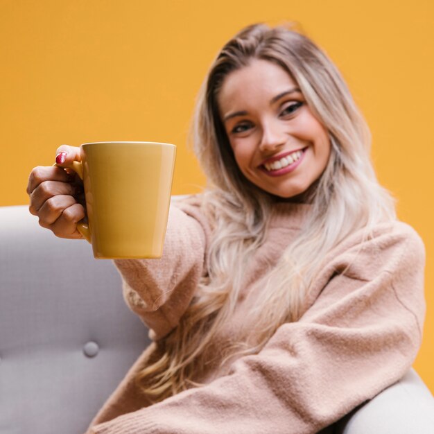 Donna felice che si siede sul sofà che mostra tazza di caffè a casa