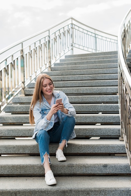 Счастливая женщина сидит на лестницах и с помощью смартфона