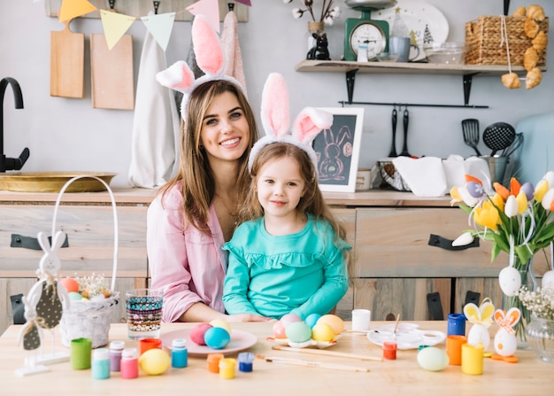 Happy woman sitting with daughter in bunny ears near Easter eggs