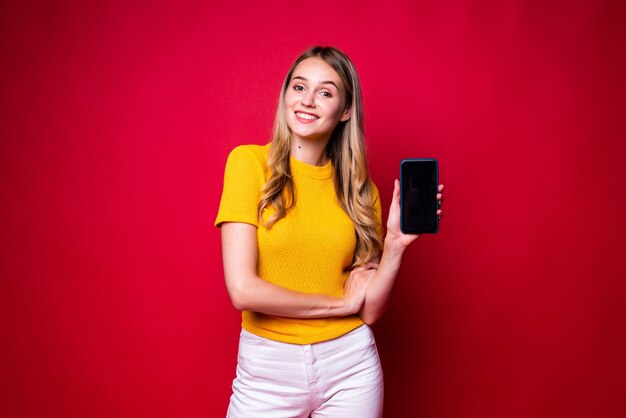 Счастливая женщина улыбается и показывает пальцем в сторону на черном экране мобильного телефона, изолированном над красной стеной