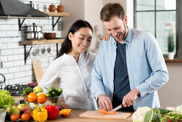 Happy woman standing near her husband slicing carrot on wooden chopping board