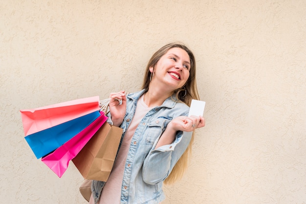 Happy woman standing with shopping bags and credit card at wall