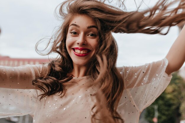 Happy woman in stylish outfit smiles widely and poses on balcony Cheerful girl in white dress with sparkles takes selfie