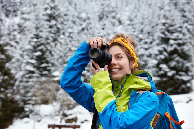 Happy woman takes photo of mountains covered with snow, spends winter holidays in nature, wears bright jacket