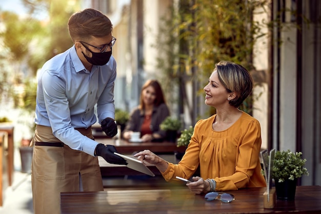 Happy woman talking to a waiter who is wearing protective face mask while choosing something from a menu on touchpad in a cafe