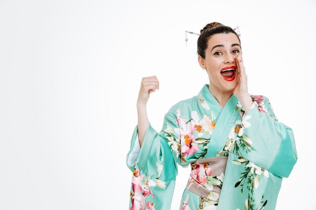 Happy Woman in traditional japanese kimono telling a secret holding hand on mouth pointing with thumb to the side being surprised on white