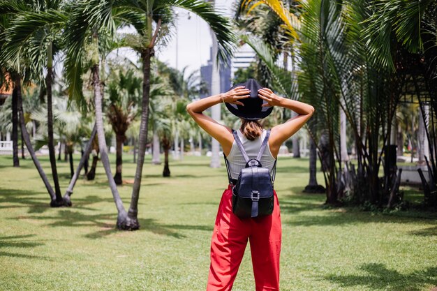 Happy woman travel around Bangkok with backpack, enjoying beautiful sunny day in in tropical park on green grass field