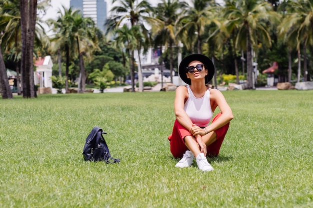 緑の芝生の上の熱帯公園で美しい晴れた日を楽しんで、バックパックを持ってバンコクを旅する幸せな女性