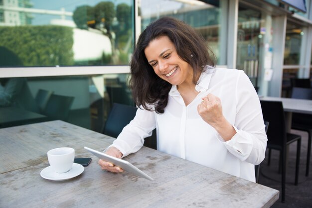 Happy woman using tablet and celebrating success in outdoor cafe