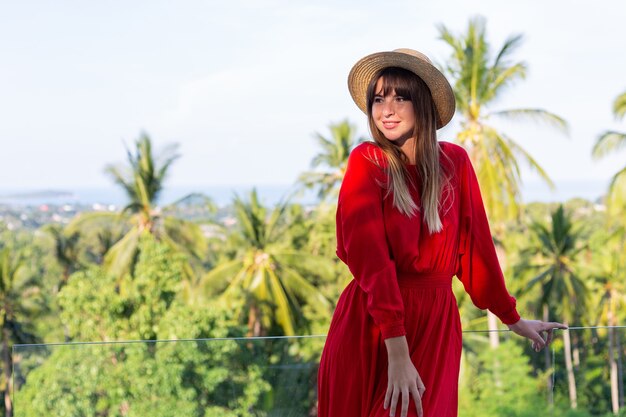 Happy woman on vacation in red summer dress and straw hat on balcony with tropical view on sea and plam trees.