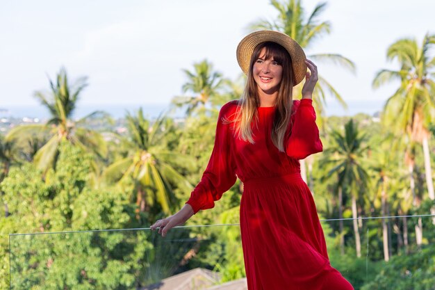 Happy woman on vacation in red summer dress and straw hat on balcony with tropical view on sea and plam trees.
