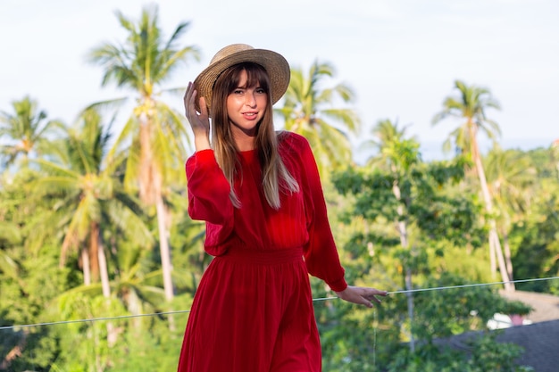 Happy woman on vacation in red summer dress and straw hat on balcony with tropical view on sea and plam trees.