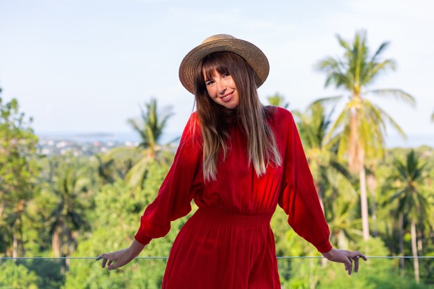 Happy woman on vacation in red summer dress and straw hat on balcony with tropical view on sea and plam trees.