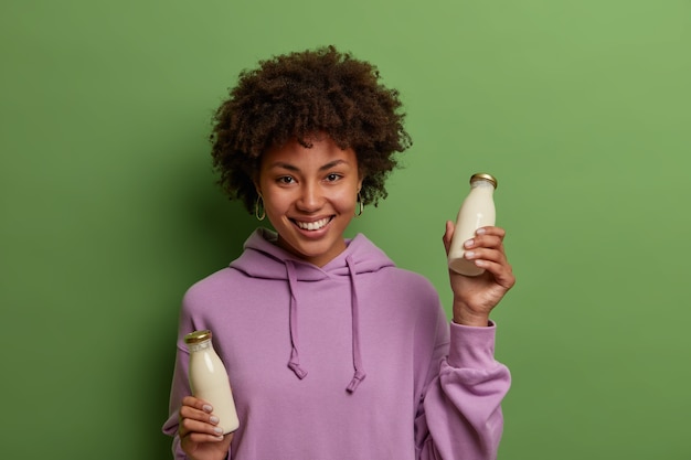Счастливая женщина-веган держит стеклянные бутылки с растительным безлактозным молоком, имеет здоровое питание, носит фиолетовую толстовку с капюшоном, широко улыбается, изолирована от зеленой стены. Немолочный альтернативный напиток
