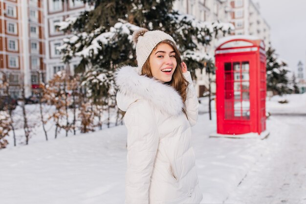 笑顔で冬の晴れた朝に歩き回って幸せな女。赤い電話ボックスと雪に覆われた通りでポーズの肩越しに見ているニット帽子の魅力的な女性