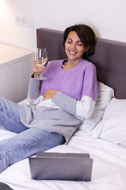 Счастливая женщина в теплом пуловере в постели с бокалом вина одна отдыхает, смотря комедию, смех фильма