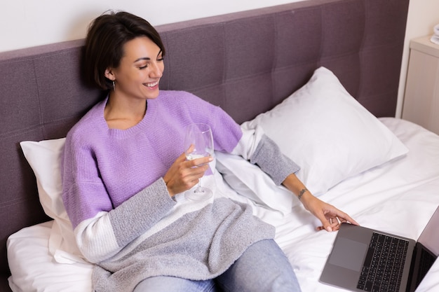 Foto gratuita donna felice in pullover caldo a letto con un bicchiere di vino da sola da sola che riposa guardando la risata di sorriso di commedia di film