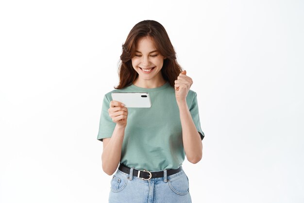 휴대폰을 보고 있는 행복한 여성과 흰색 배경에 스마트폰으로 서서 승리를 축하하는 온라인 승리를 기뻐하는 여성