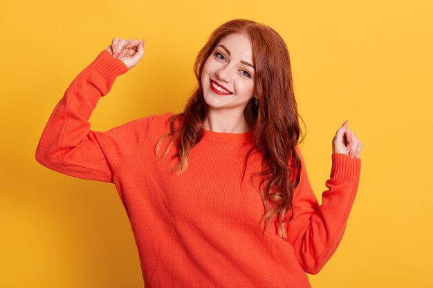 Happy woman wearing orange sweater, looking smiling at camera with raising hands, standing isolated