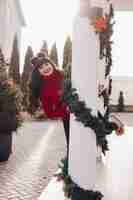 Free photo happy woman wearing red sweater and peeking from behind a white column decorated with tinsel