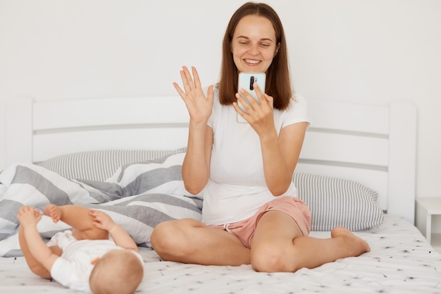 Happy woman wearing white t shirt and shorts having video call or broadcasting livestream, waving hand to device camera and smiling, posing while sitting on bed at home with little baby.