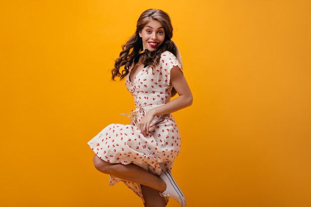 Happy woman in white dress jumps on orange background.Beautiful stylish girl with curly hair in summer outfit and light sneakers smiling.