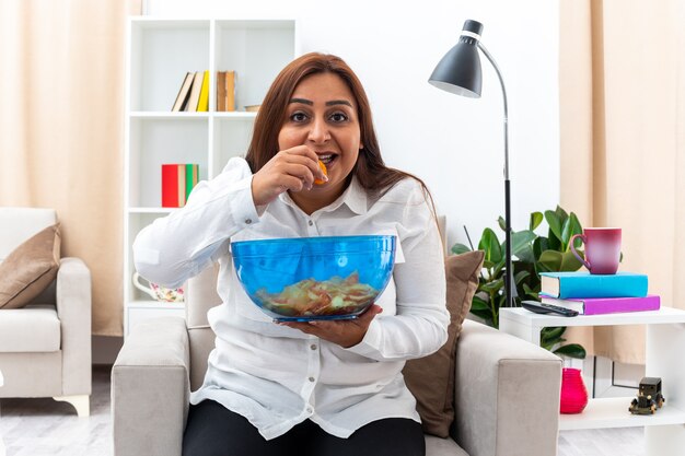 Happy woman in white shirt and black pants relaxing sitting on the chair with bowl of chips eating chips in light living room