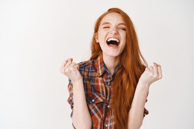 Happy woman winning scream of joy and triumph Redhead girl achieve goal and celebrating shouting for rejoice standing over white background