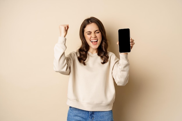 Happy woman winning showing smartphone screen and shouting of joy fist pump standing over beige back...