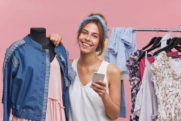 Happy woman with appealing appearance, standing near mannequin, having joyful expression while being glad to buy new outfit, typing messages to her best friend, sharing latest news with each other