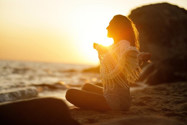 腕を伸ばして砂浜でリラックスして夏の夕日を楽しむ幸せな女性
