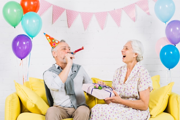 Happy woman with birthday gift looking at her husband blowing party horn