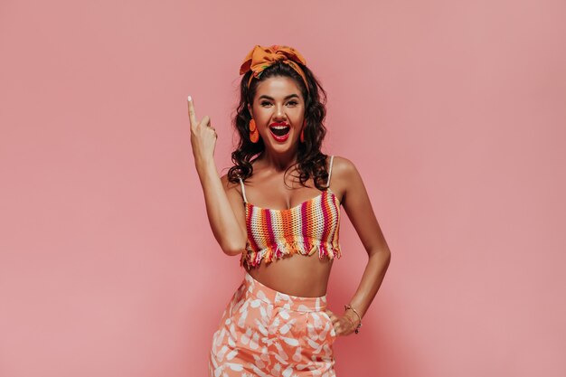 Happy woman with black curly hairstyle and orange headband in modern top and pink cool pants showing up and looking into camera..