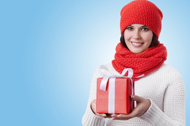 明るい茶色の目、黒い髪、赤いスカーフ、帽子を身に着けている彼女の手でプレゼントを持っている魅力的な笑顔で幸せな女