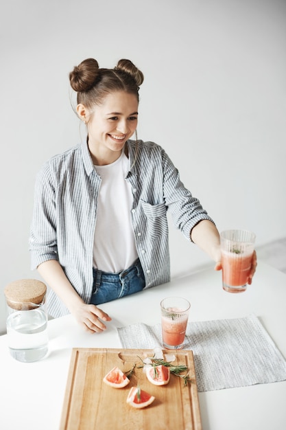 Happy woman with buns smiling stretching glass with grapefruit detox smoothie to somebody. White wall background. Healthy diet food.
