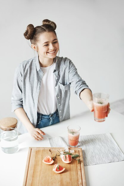 Happy woman with buns smiling stretching glass with grapefruit detox smoothie to somebody. White wall background. Healthy diet food.