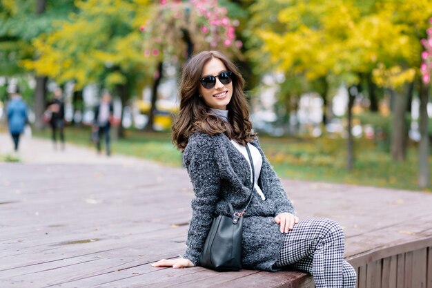 Happy woman with charming smile and dark hair relaxes in the autumn park in sunlight. She is sitting on the wooden table and laughing in the park with yellow trees and bushes.