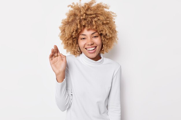 巻き毛のふさふさした髪の幸せな女性は、手を上げて笑顔を保ち、前向きな感情を誠実に表現します。白い背景の上に隔離されたカジュアルなタートルネックを着て、何か面白いものを聞きます。幸福の概念