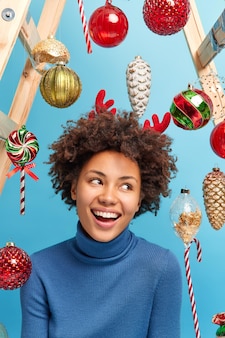 La donna felice con i capelli ricci distoglie lo sguardo sorride felice a trentadue denti andando a festeggiare il nuovo anno decora la stanza prima che l'evento festivo si ponga contro i giocattoli isolati su sfondo blu. decorazione natalizia