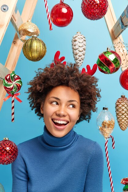 Happy woman with curly hair looks away gladfully smiles toothily going to celebrate New Year decorates room before festive event poses against toys isolated over blue background. Holiday decoration