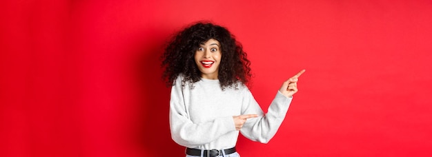 Free photo happy woman with curly hair smiling amused pointing fingers left at logo and looking amazed checking