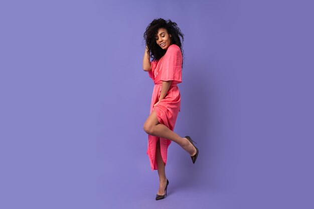Счастливая женщина с вьющимися волосами, позирует над фиолетовой стеной. Носить элегантное вечернее платье. Весенний модный образ. Полная длина.