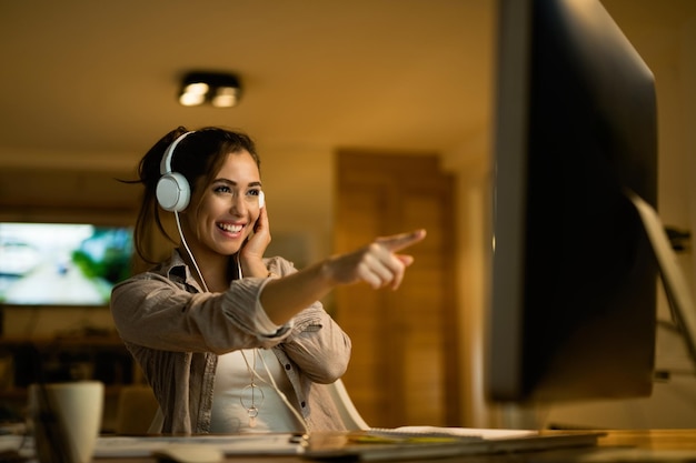 Foto gratuita donna felice con le cuffie che puntano allo schermo del computer mentre navighi in rete la sera a casa