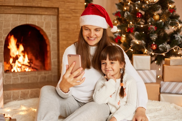 Счастливая женщина с ее милой маленькой дочерью сидит на полу возле елки и камина, держит смартфон, смотрит на экран устройства, имеет позитивные выражения лица и праздничное настроение.