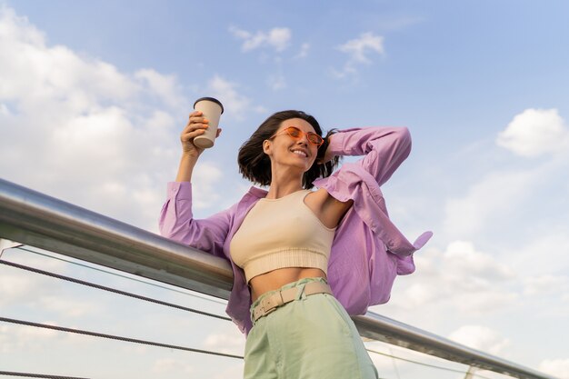 현대 다리를 걷는 동안 커피 한잔 즐기는 세련된 보라색 특대 셔츠에 완벽한 그림으로 행복한 여자