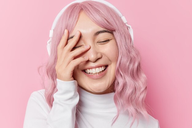 Happy woman with pink dyed hair covers face with hands feels positive keeps eyes closed smiles toohily wears casual white jumper isolated over rosy background. Emotions and lifestyle concept