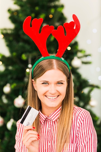 Happy woman with plastic card in deer antlers headband near Christmas tree