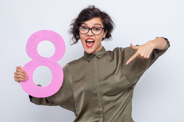 Happy woman with short hair holding number eight made from cardboard pointing with index finger down smiling cheerfully celebrating international women's day march 8 standing over white background