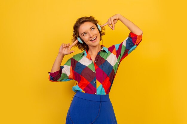 Happy woman with short hairstyle listening to music by earphones and having fun over yellow wall