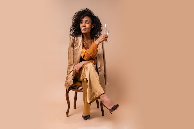 Happy  woman with tan skin with perfect curly hairs  holding glass of wine ,wearing ielegant orange blouse and silk pants sitting on vintage chair  beige wall.